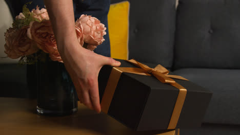 Nahaufnahme-Eines-Mannes,-Der-Ein-Als-Geschenk-Verpacktes-Geschenk-Mit-Einer-Dankeskarte-Und-Blumen-Auf-Den-Tisch-Im-Wohnzimmer-Zu-Hause-Legt